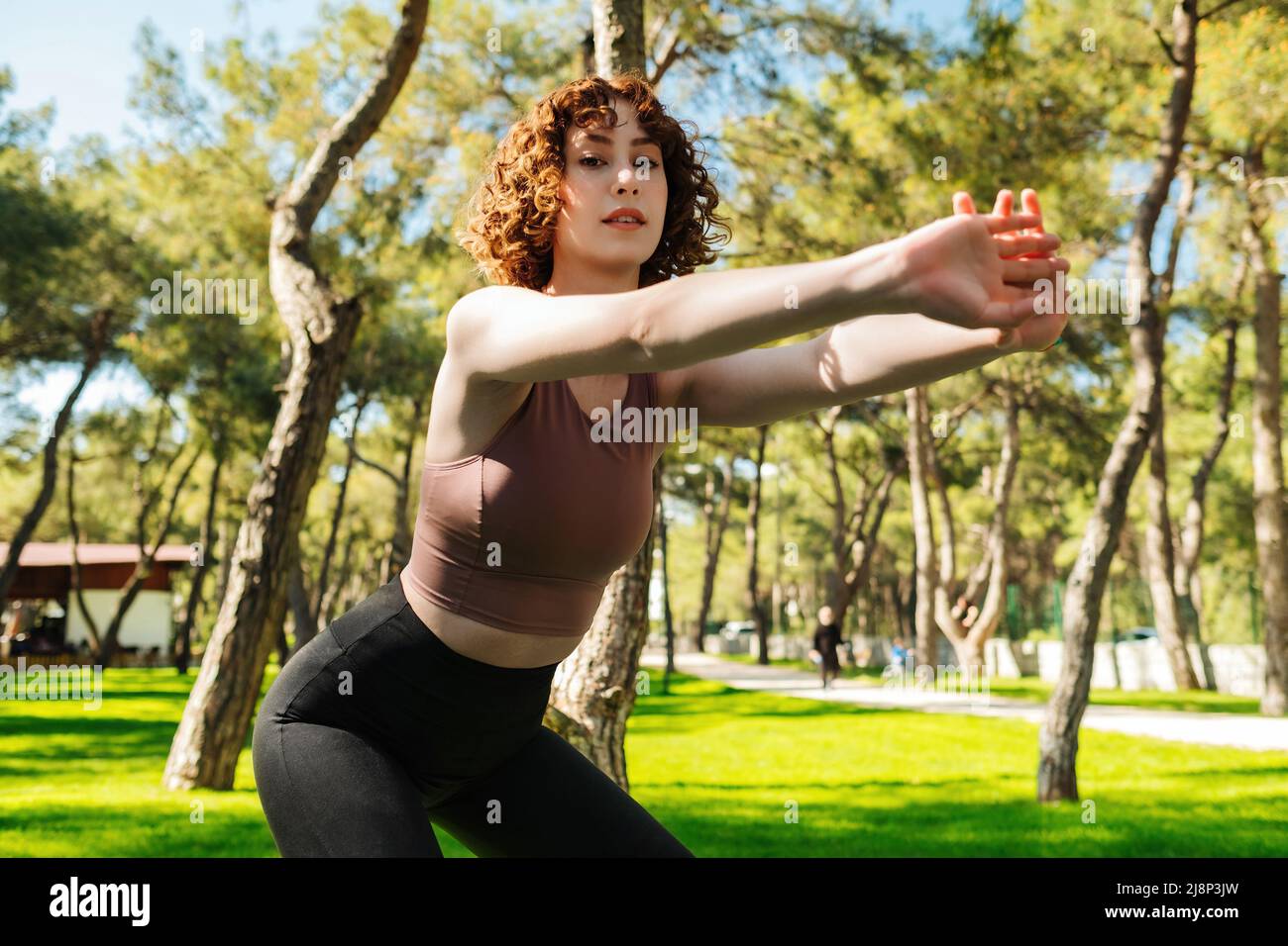 Una giovane donna rossa che dura nel tempo si allena al parco cittadino, all'aperto facendo squat profondi con le braccia distese. Donna sportiva che guarda la fotocamera. Sport c Foto Stock