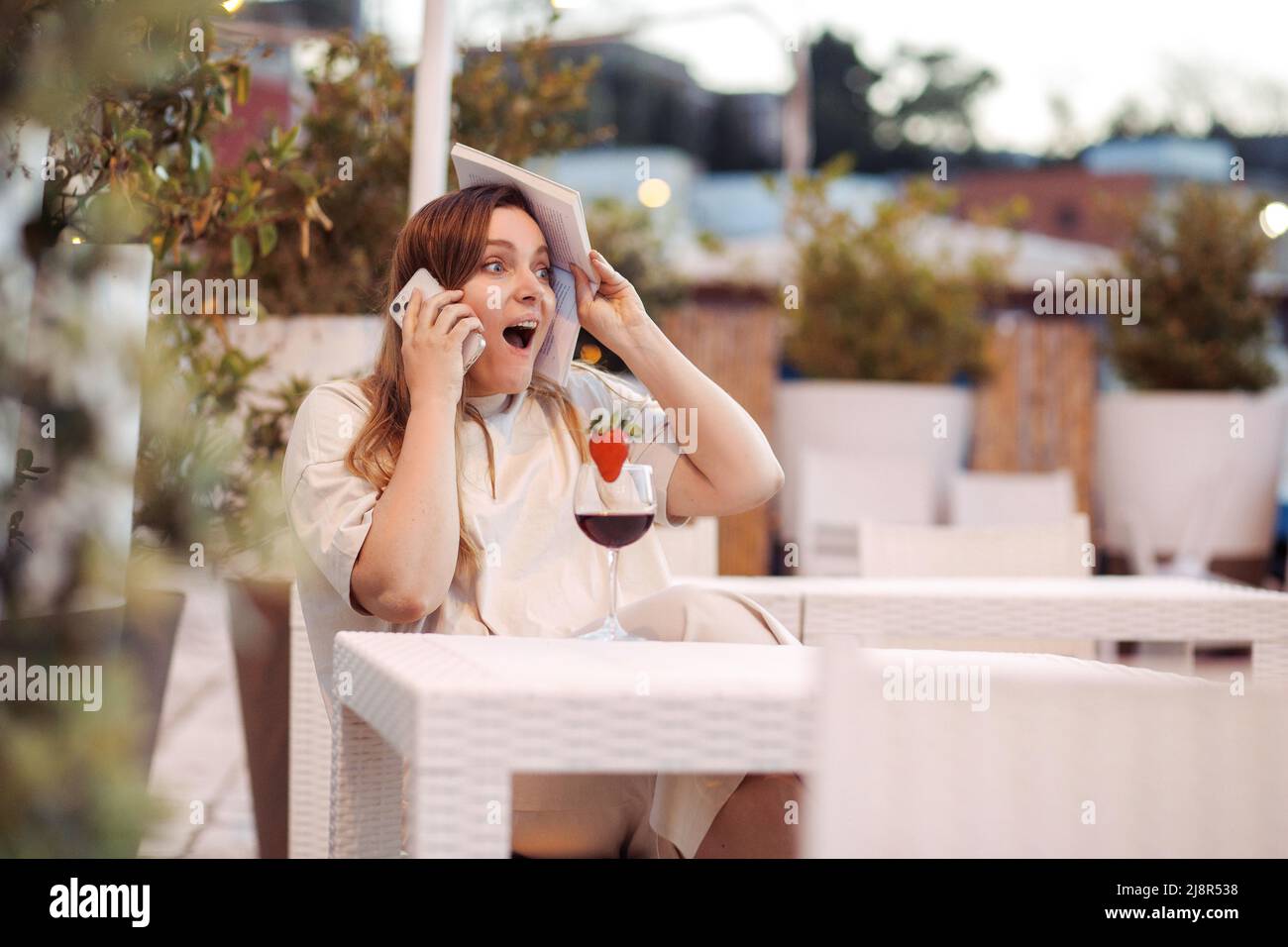 Affascinante donna esclamante con libro in mani sedersi al tavolo con drink in ristorante ottenere buone notizie sul telefono cellulare Foto Stock