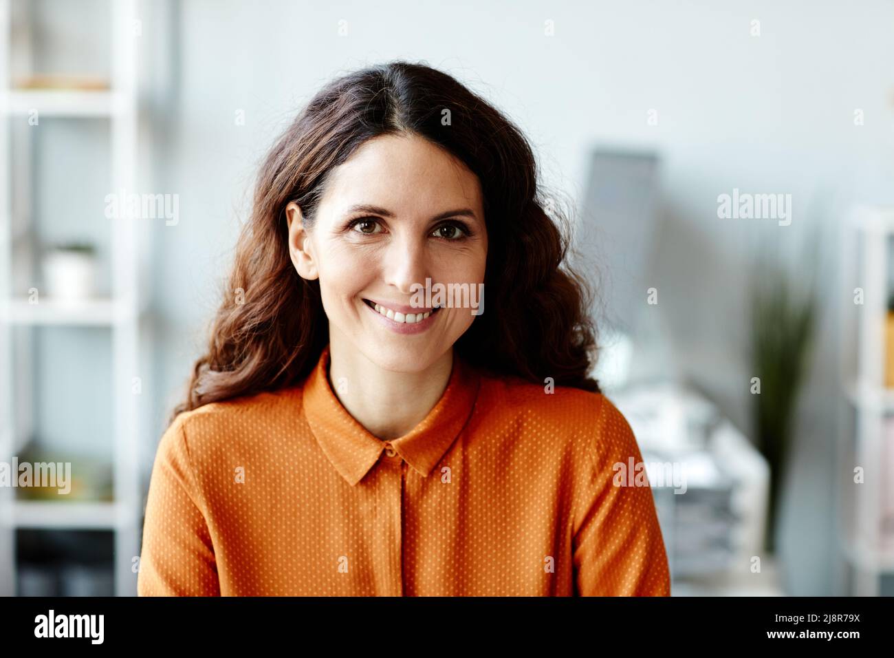 Medio primo piano di affascinante donna caucasica con lungo marrone scuro sentire seduto in ufficio sorridendo alla macchina fotografica Foto Stock