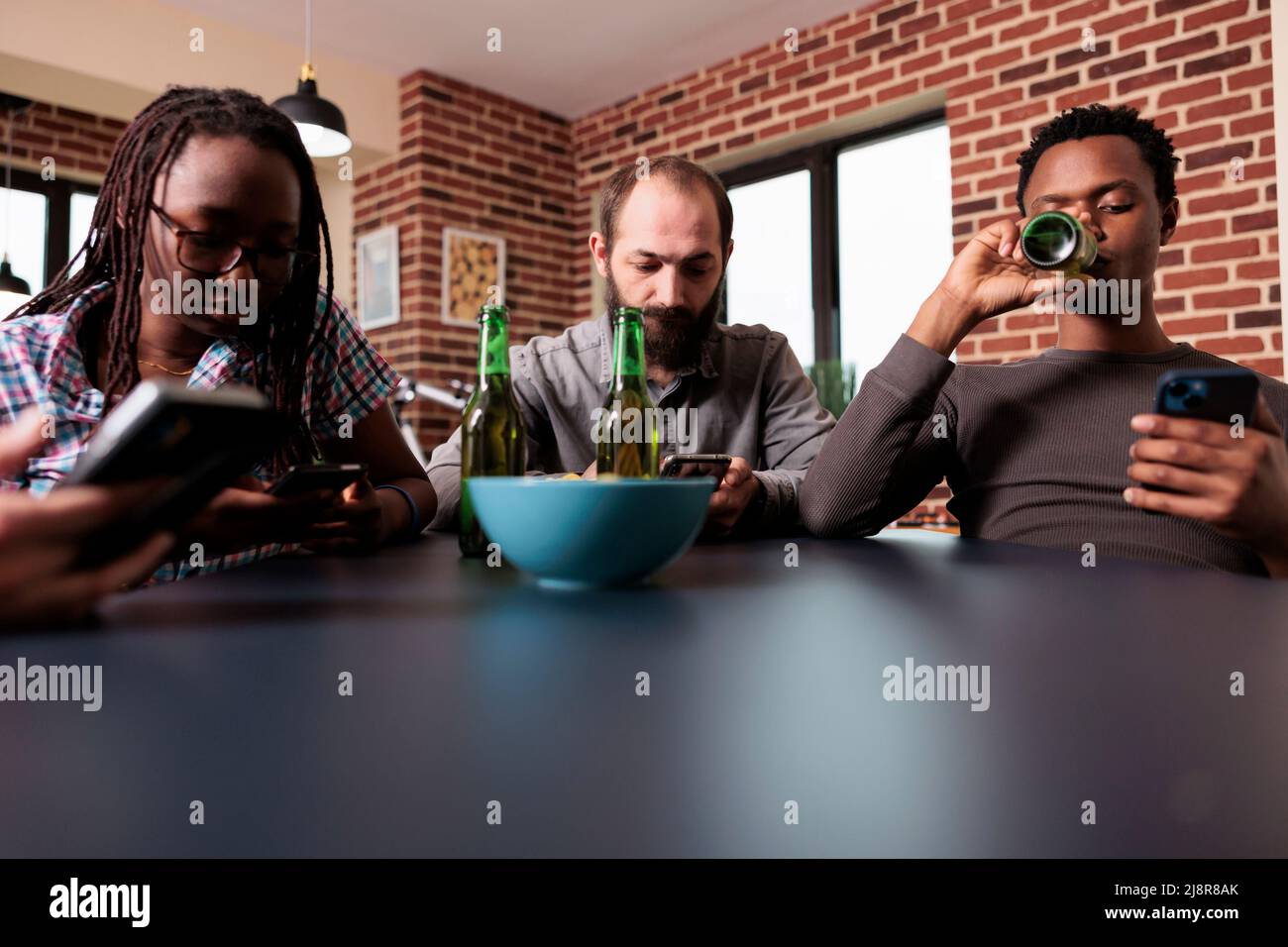Gruppo diversificato di amici seduti in soggiorno con smartphone. Persone multirazziali a casa navigando pagine web su moderni dispositivi elettronici touchscreen mentre si hanno birra e snack. Foto Stock