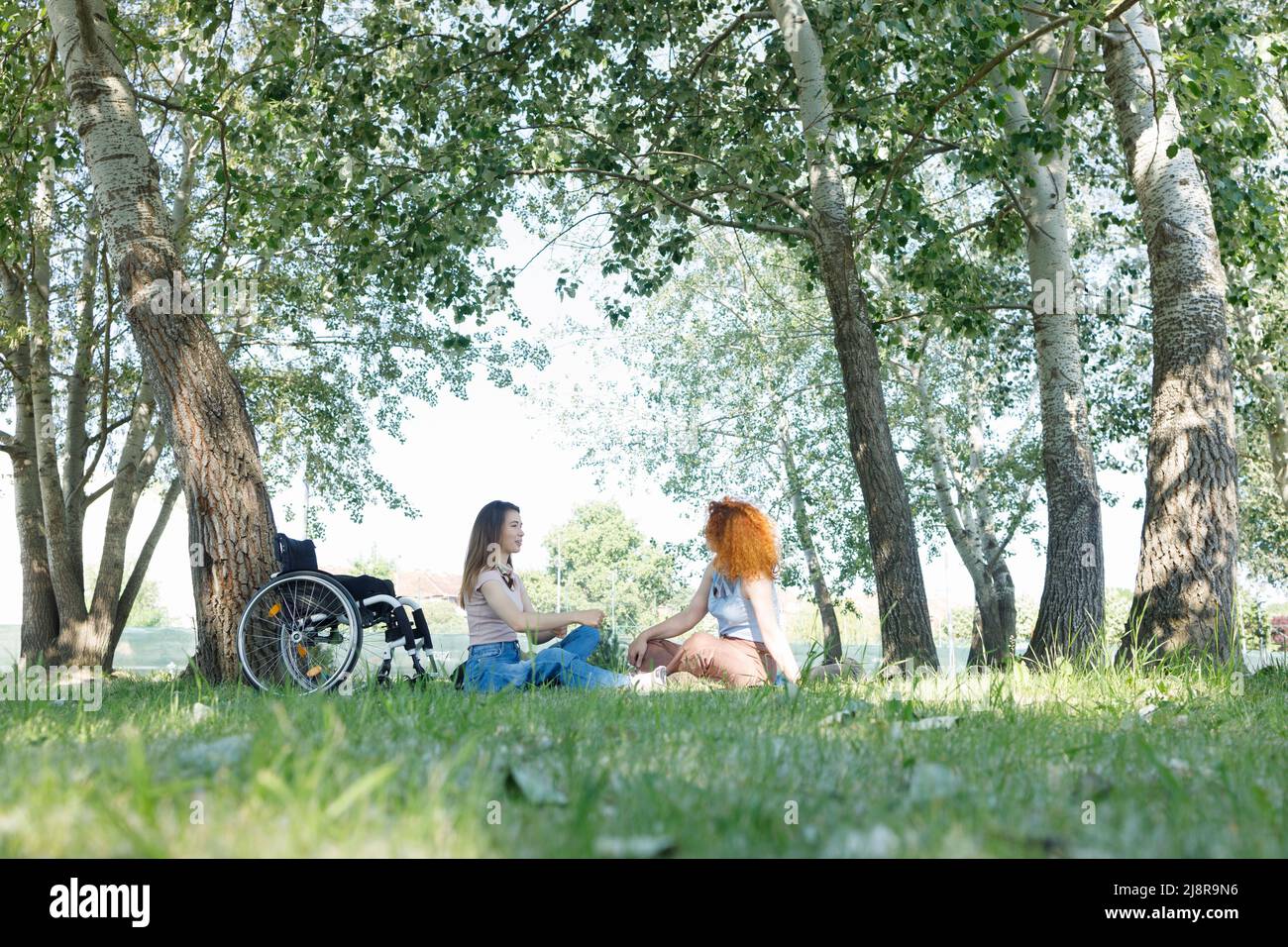 Una donna con disabilità e la sua amica femmina che ha una conversazione all'ombra di splendidi alberi di pioppo, durante una piacevole giornata estiva Foto Stock
