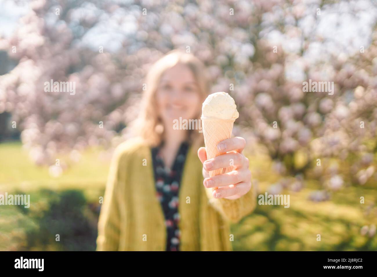 Giovane sorriso 20s donna che si sente felice, dando un grande gelato alla vaniglia in cono waffle su sfondo albero fiorente. Concetto estivo Foto Stock