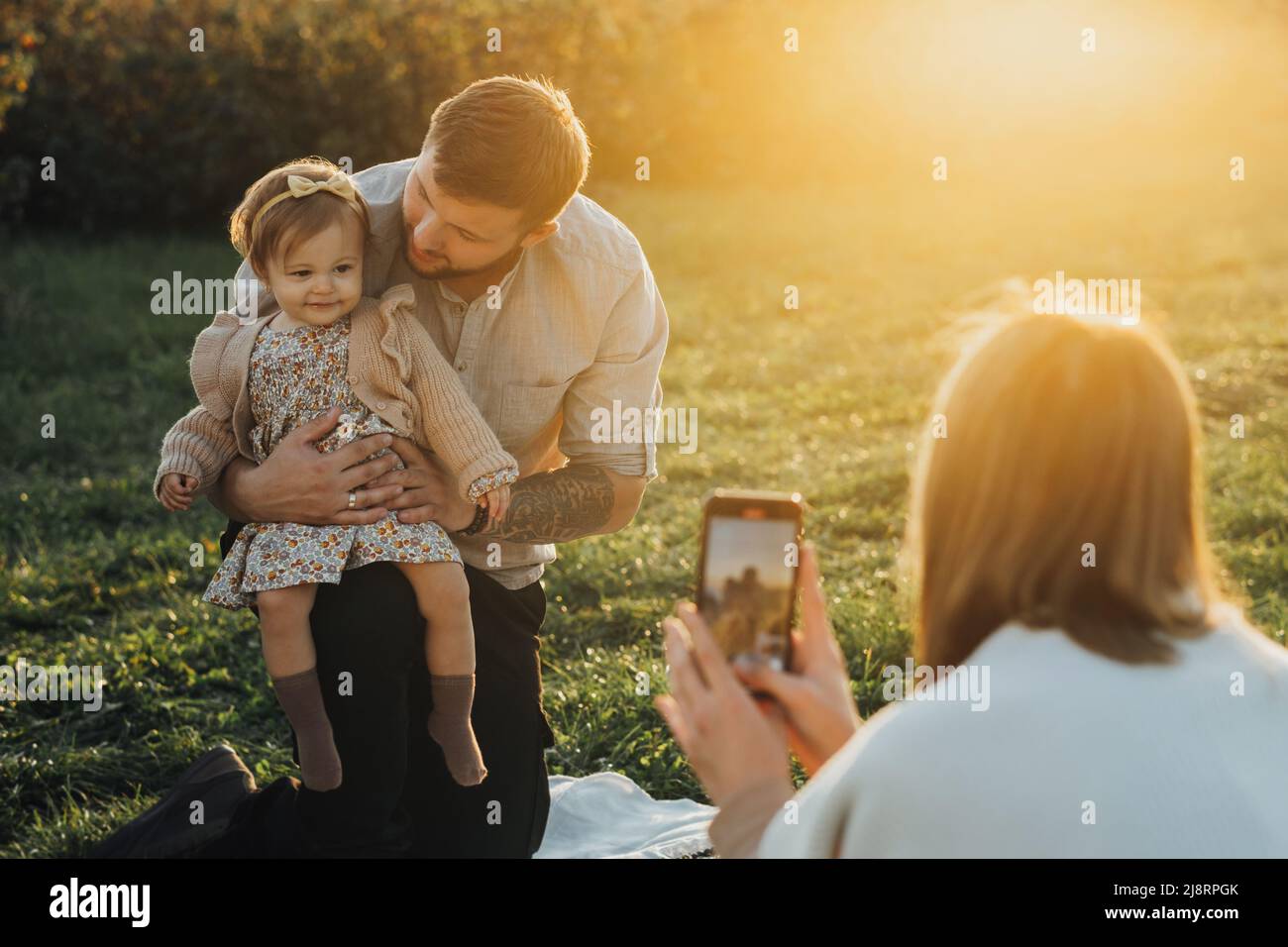 Felice giovane famiglia che ha picnic all'aperto, donna che prende le immagini del suo marito e della figlia del bambino al tramonto Foto Stock