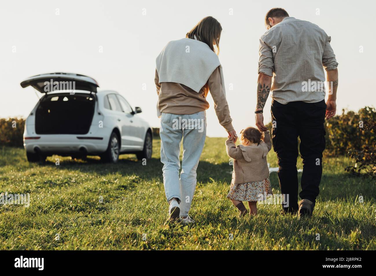 La giovane famiglia si diverta a fare un viaggio su un'auto SUV, madre e padre tatuato aiutando la figlia del bambino a fare i primi passi all'aperto sul campo al tramonto Foto Stock