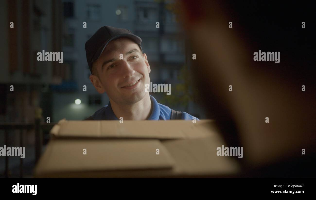 Il corriere giovane attraente di consegna in blu sorridente nel cliente sta trasportando le scatole intorno. Lavoratore urbano. Foto Stock
