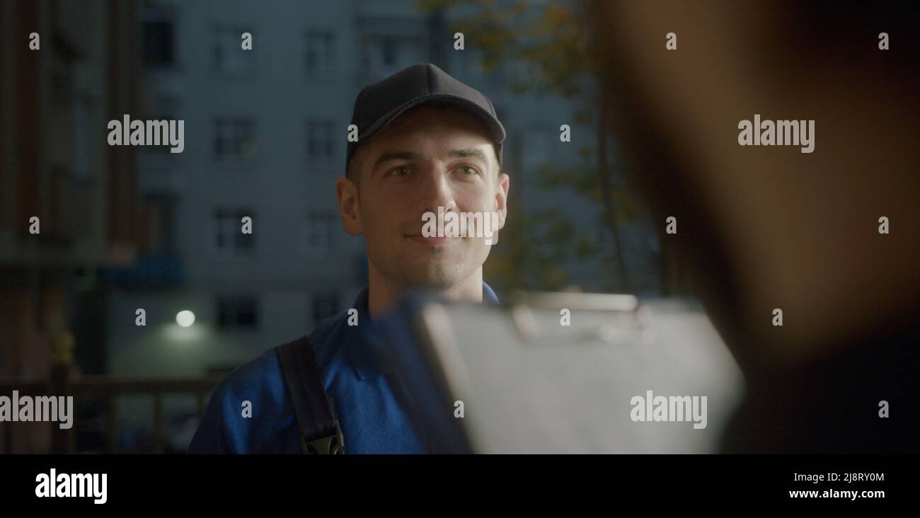 Attraente giovane corriere di consegna in blu cappuccio nel cliente che gli dà una bolla di consegna per firmare. Lavoratore urbano Foto Stock