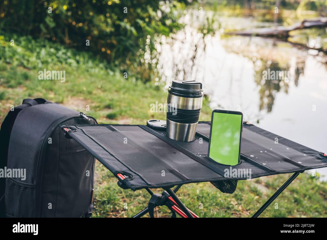Tavolo da campeggio con thermo Cup e smartphone all'aperto. Schermo verde. Paesaggio pittoresco. Concetto di svago e di viaggio. Tecnologia Foto Stock