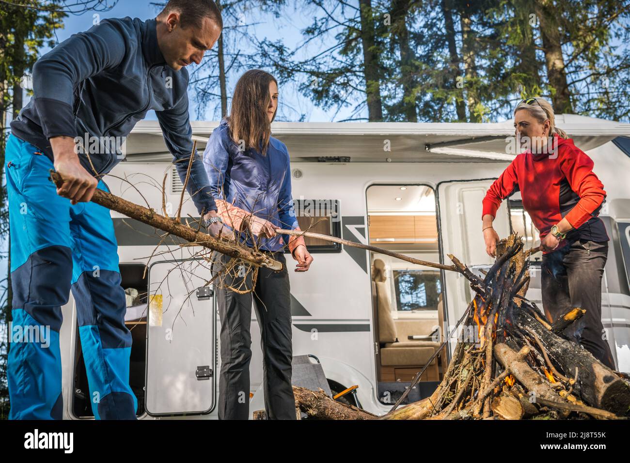 Tre amici caucasici che si aggirano intorno a Campfire accanto al loro veicolo ricreativo Motorhome. Tema Getaway weekend estivo. Foto Stock