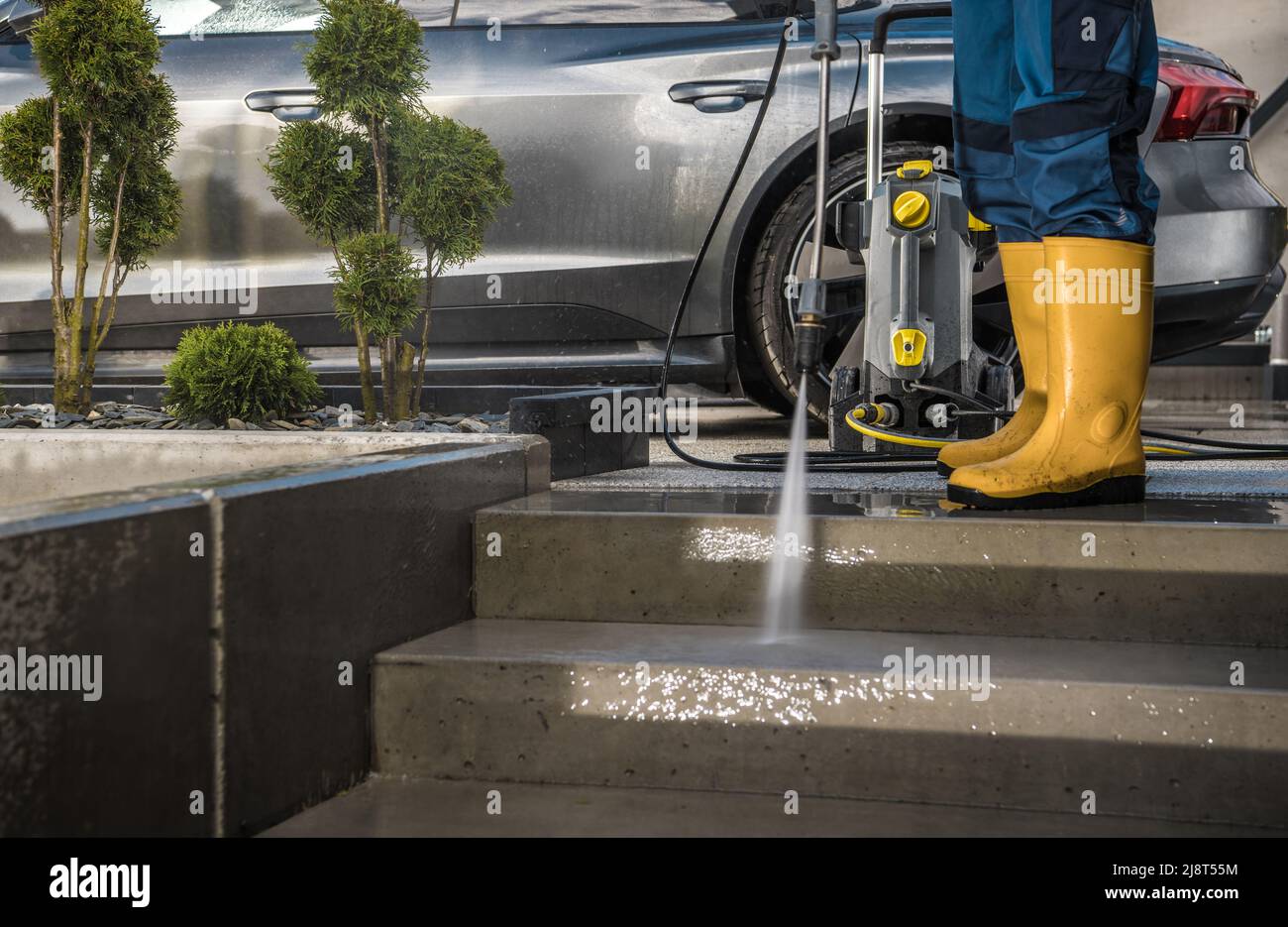 Proprietario di abitazione moderno che lava i suoi percorsi del giardino e vialetto di guida usando la rondella di pressione potente vicino alla sua automobile. Home dintorni manutenzione. Foto Stock