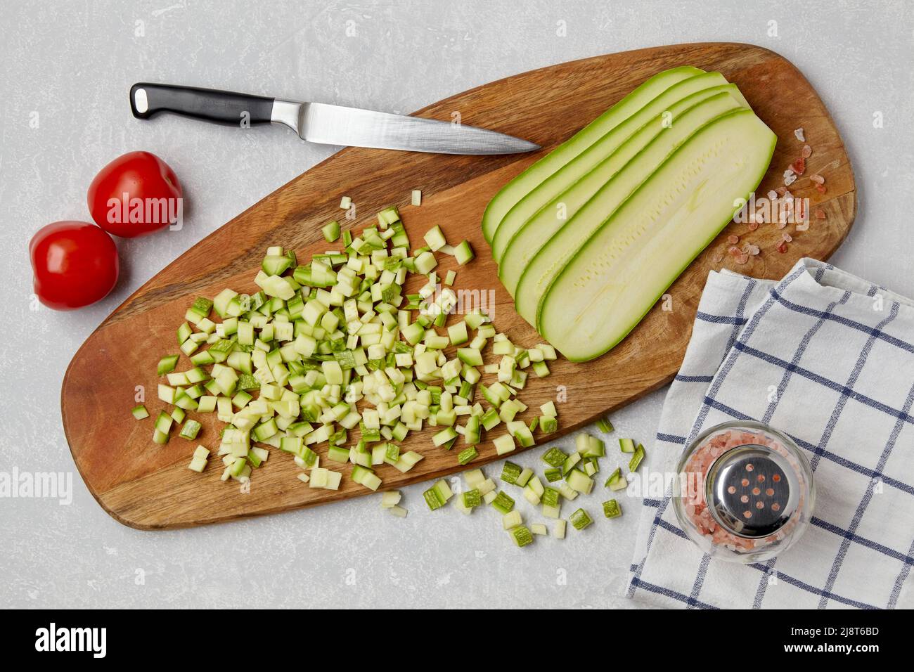 Affettare le zucchine di zucchine su un tagliere di legno, coltello, pomodori, sale rosa e tovagliolo su una tavola leggera in cemento. Vista dall'alto piatto lat Foto Stock