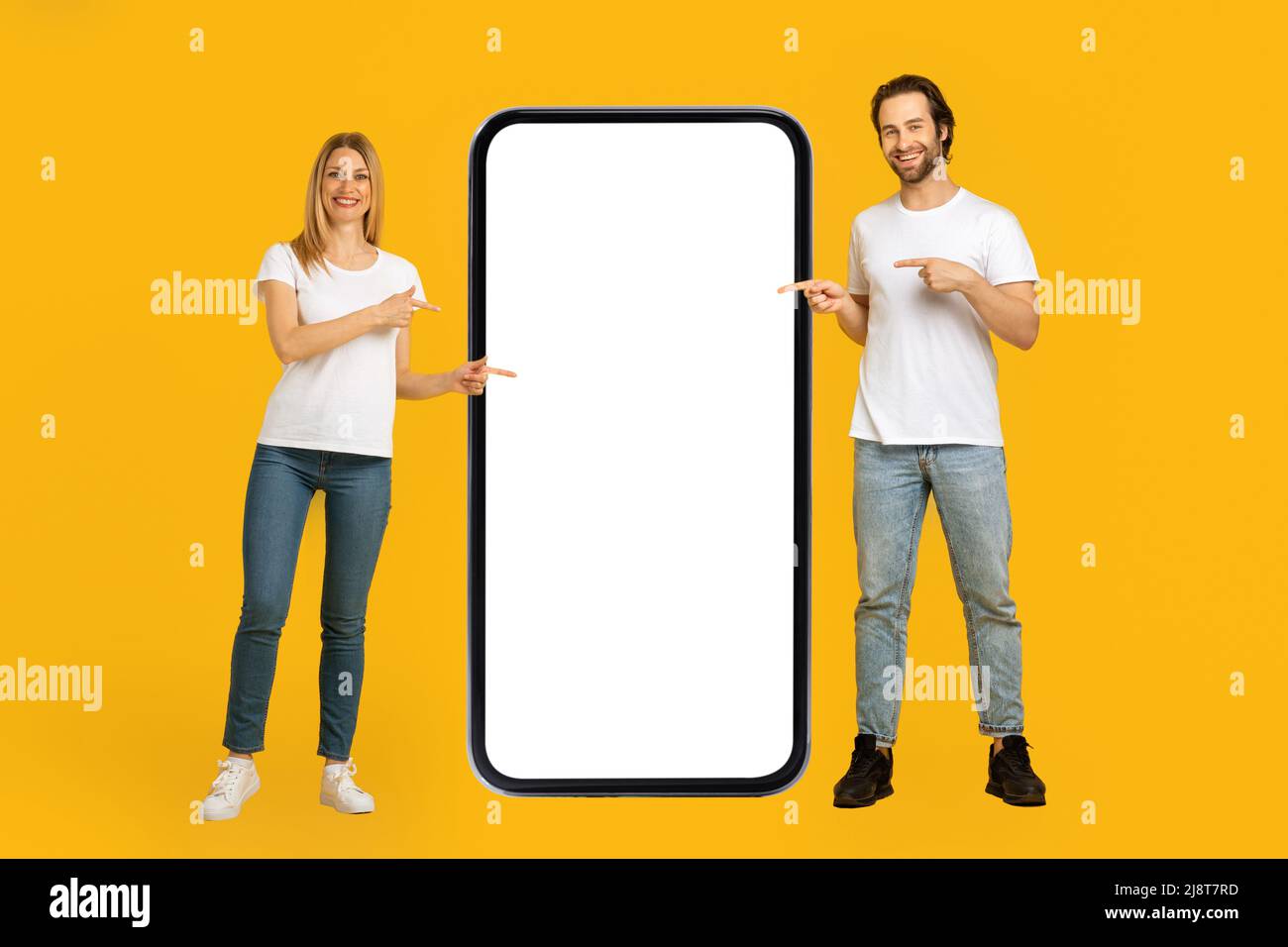 Allegro giovane coppia europea in magliette bianche puntano le dita su un telefono enorme con schermo vuoto Foto Stock