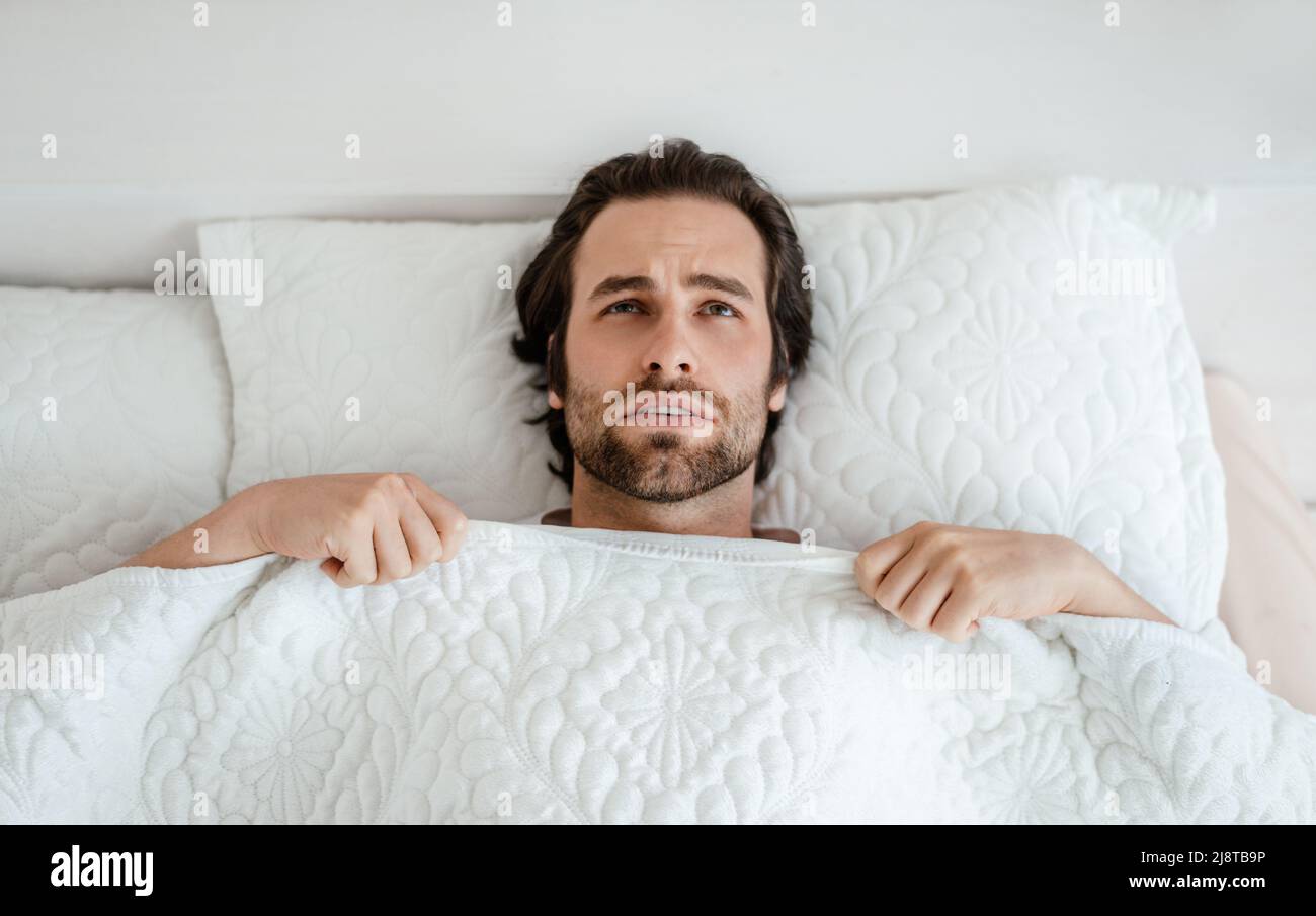 Infelice uomo caucasico millenario con sveglia stoppia, sensazione di male, sdraiato sul letto bianco in camera da letto interno Foto Stock