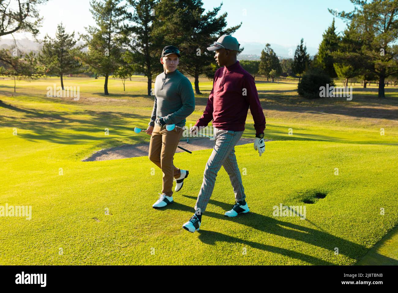 Giovani amici maschi multirazziali con club di golf che parlano mentre si cammina al campo di golf in estate Foto Stock