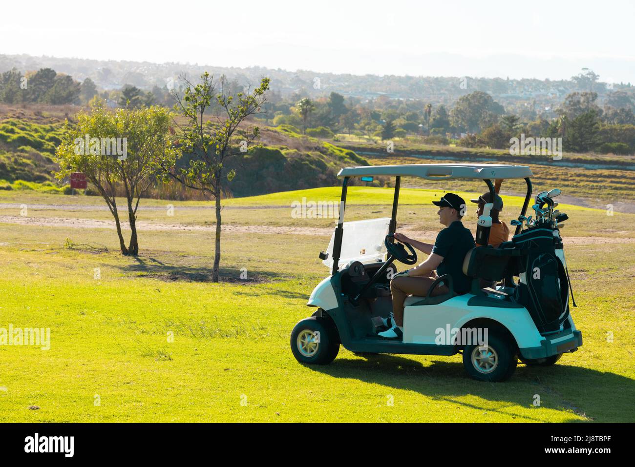 Amici giovani uomini multirazziali che cavalcano in golf cart contro il cielo limpido al campo d'estate Foto Stock