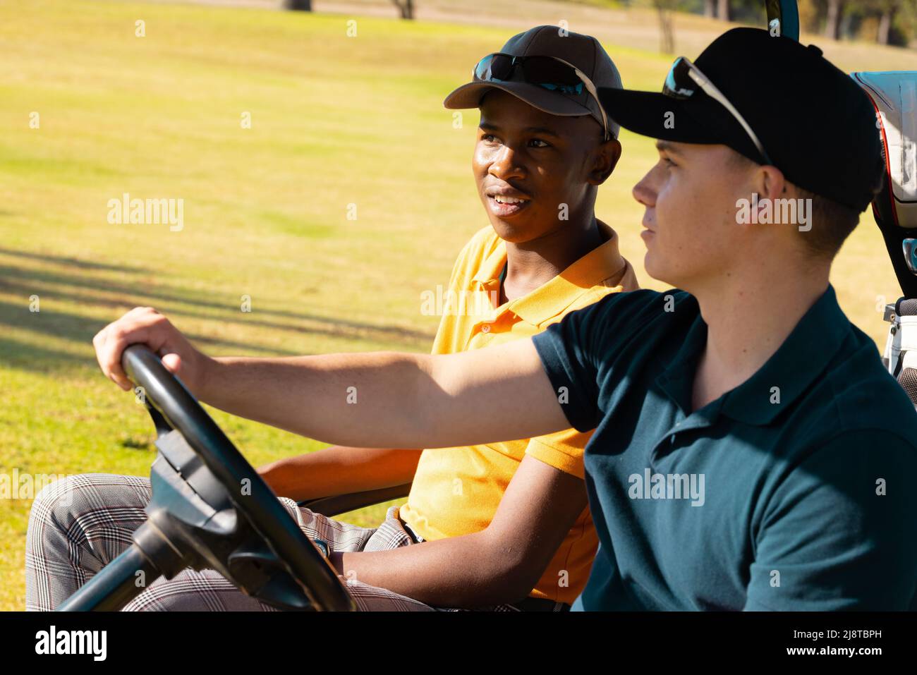 Giovane uomo caucasico e amico afroamericano che indossa tappi seduti in golf cart presso il campo da golf Foto Stock