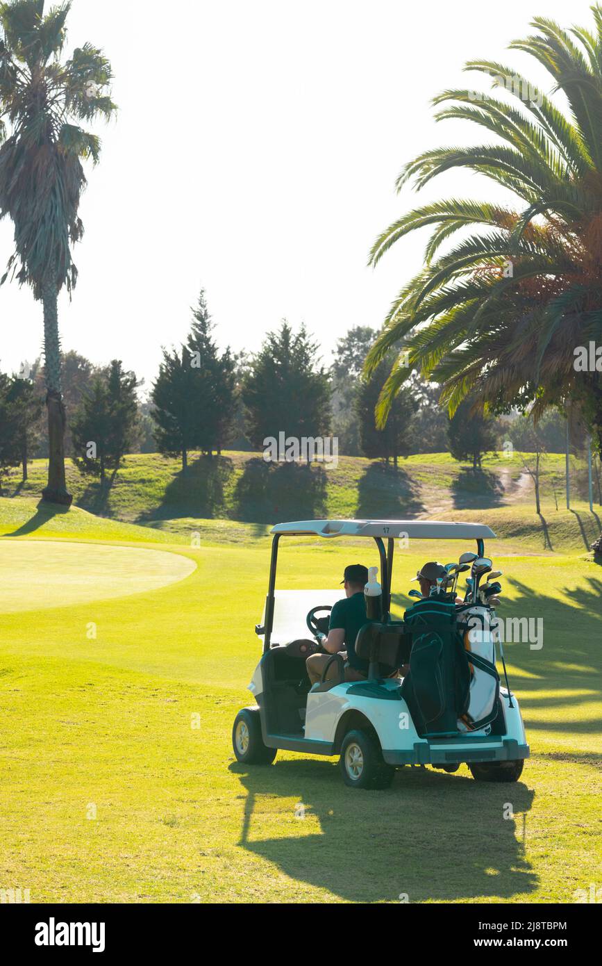 Amici maschi giovani multirazziali con borse seduti in golf cart contro il cielo limpido al campo da golf Foto Stock