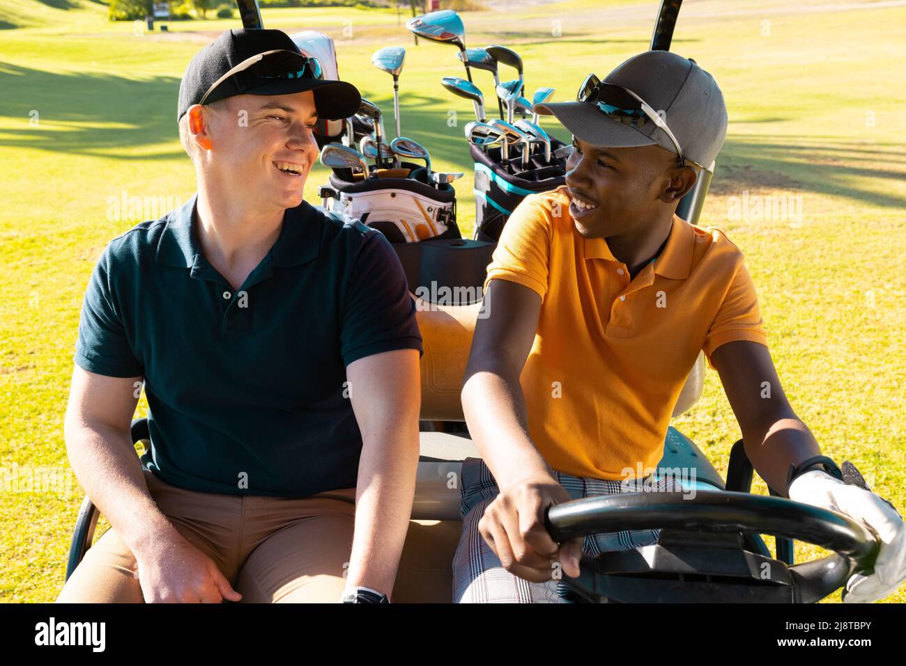 Felice multirazziale giovani amici maschi che indossano tappi parlando mentre si siede in golf cart al campo da golf Foto Stock