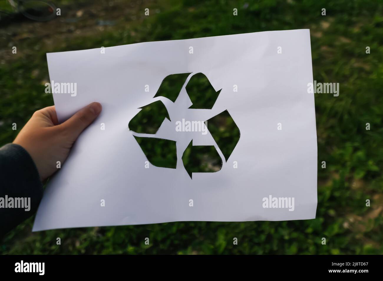 Defocus mano tenendo carta tagliata con il logo di verde erba natura spazzatura sfondo. Concetto di riciclaggio. Inquinamento del pianeta. Fuori fuoco. Foto Stock