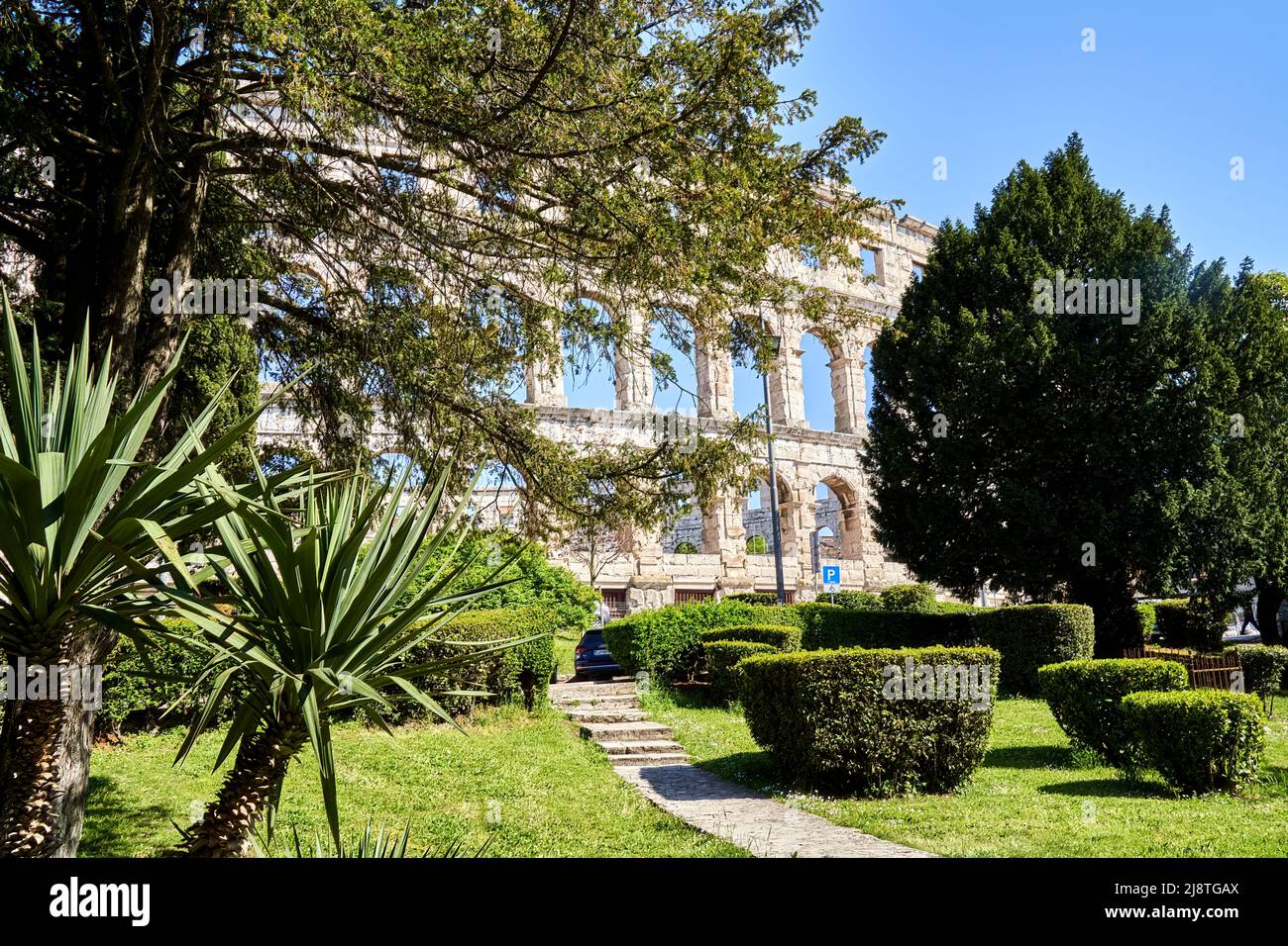 Parco con prato ben curato, palme, ulivi, siepi e cipressi di fronte alle rovine di un anfiteatro romano Foto Stock