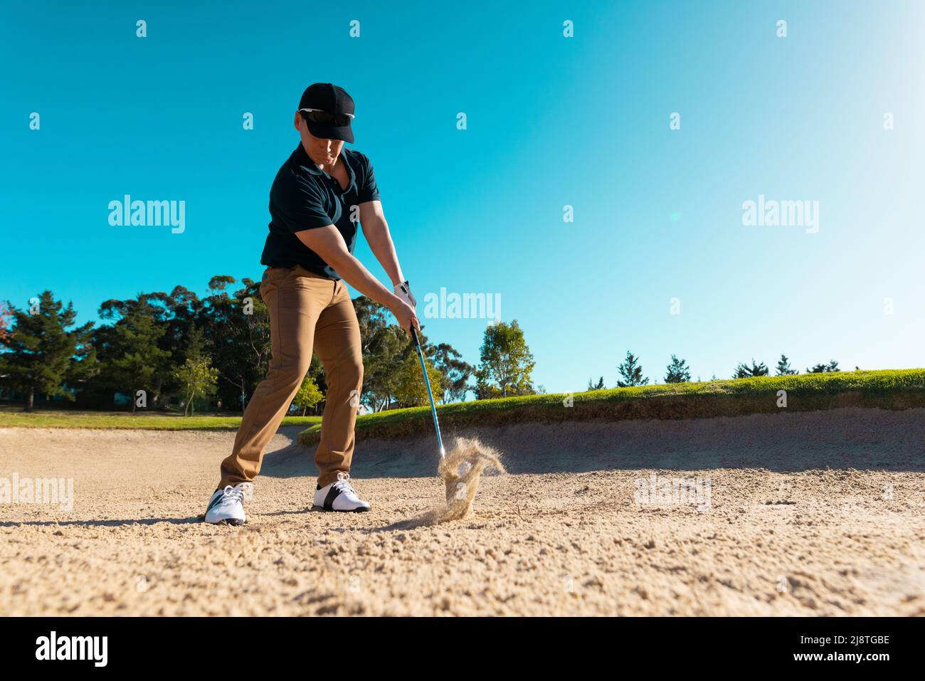 Vista ad angolo basso del giovane caucasico che colpisce con golf club su terra sabbiosa contro cielo blu chiaro Foto Stock