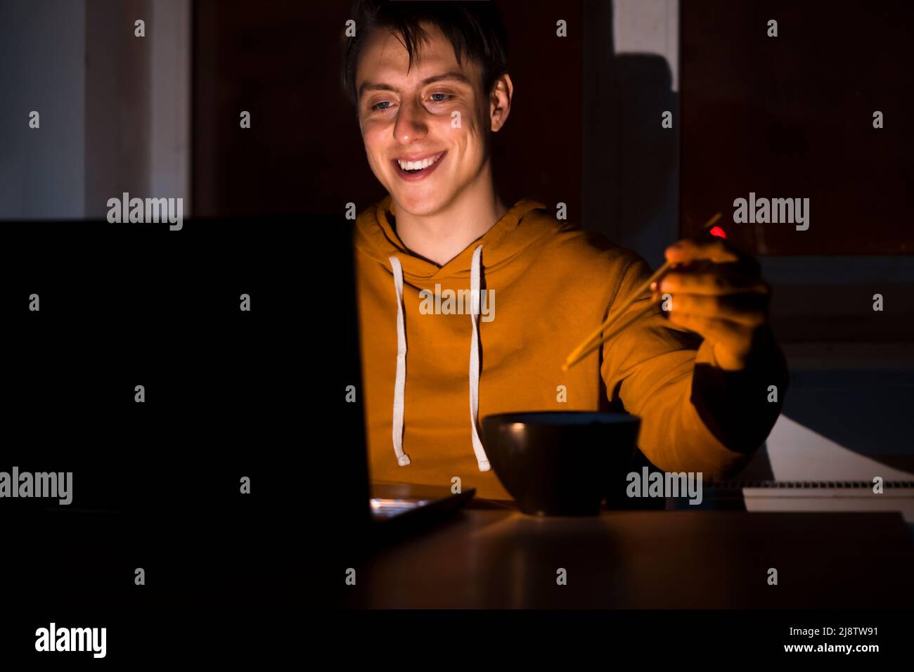 L'uomo mangia tagliatelle con bastoncini e lavora su un computer portatile. Foto Stock