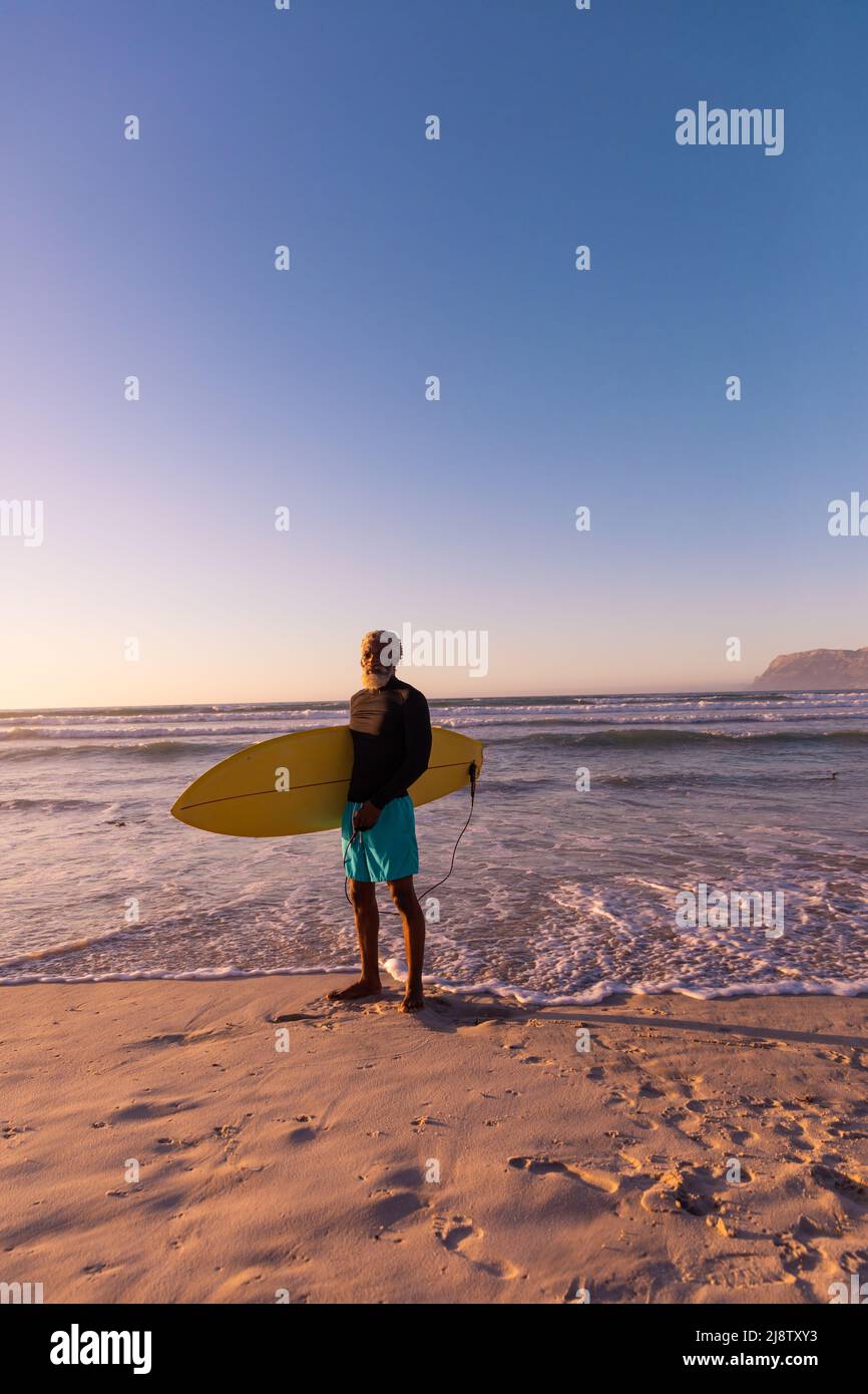 Uomo maggiore afro-americano con tavola da surf in piedi contro il mare e il cielo blu chiaro Foto Stock