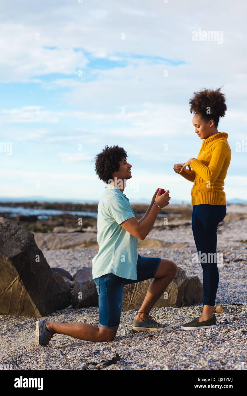 Giovane afroista con ring box che propone una ragazza afro-americana mentre si inginocchiano in spiaggia Foto Stock