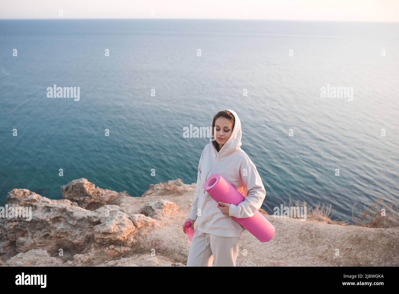 Bella donna indossare cappuccio che tiene rosa yoga tappeto in piedi in cima alla roccia sopra mare costa all'aperto. Fitness attivo della ragazza adulta. Benessere sano Foto Stock