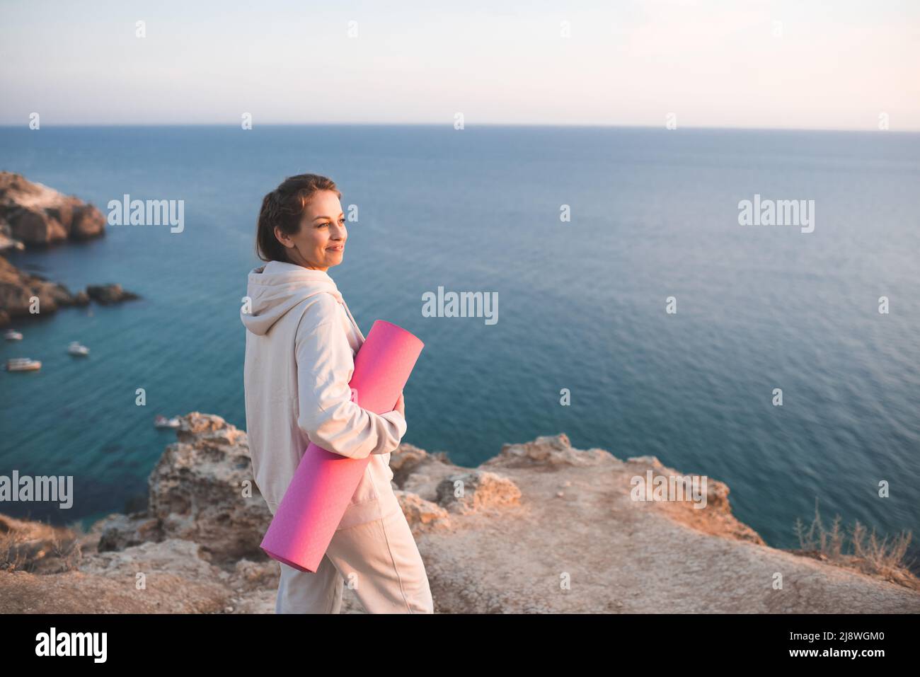 Donna sorridente felice che tiene rosa il tappeto yoga indossare felpa con cappuccio sportivo e pantaloni in piedi sulla roccia su sfondo lato mare. Practic ragazza adulta grazioso Foto Stock