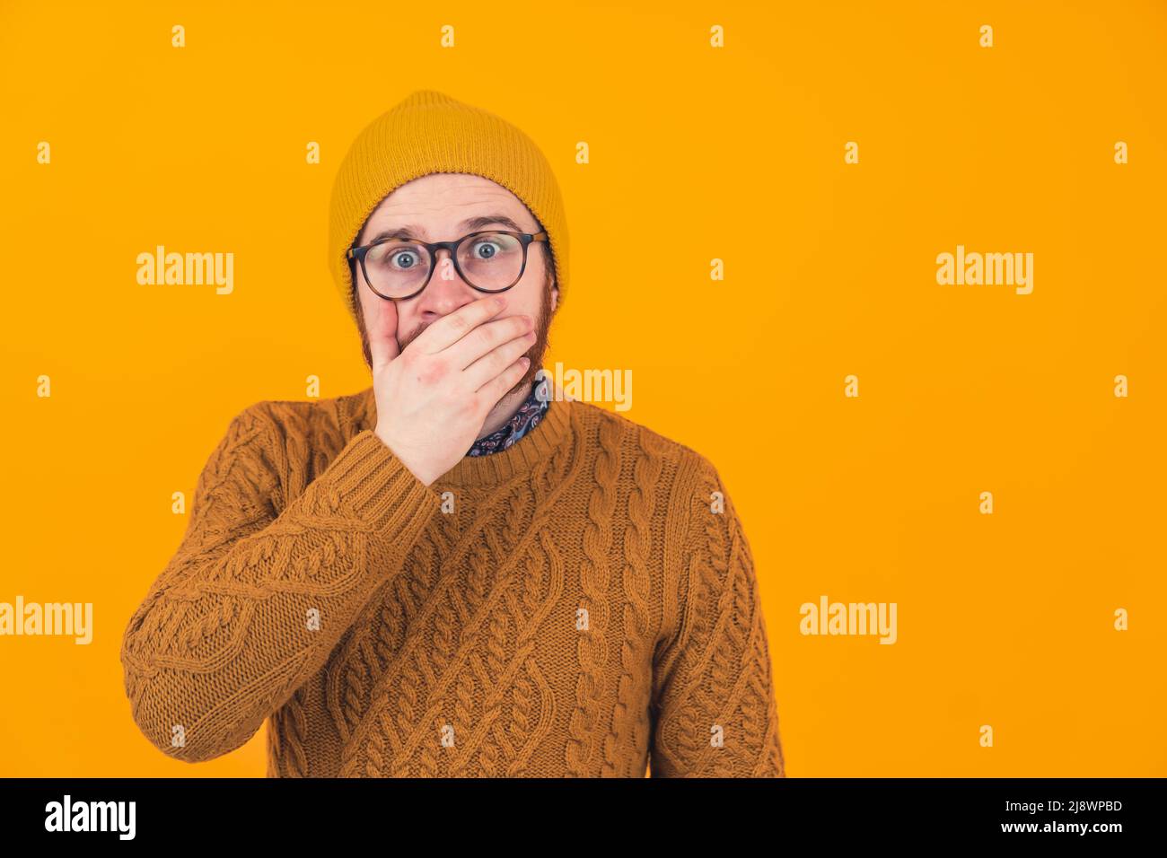 Studio girato su sfondo arancione di un isolato scioccato spaventato uomo caucasico sorpreso guardando la macchina fotografica con gli occhi spalancati coprendo la bocca con la mano che indossa un maglione e un cappello. Foto di alta qualità Foto Stock