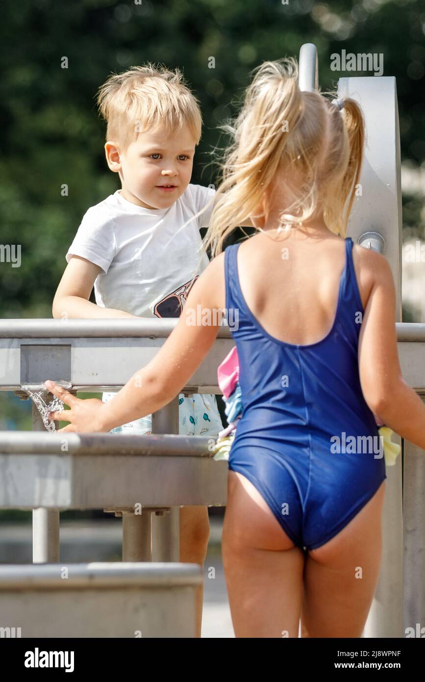 Un ragazzo felice e bella ragazza in un costume da bagno blu gioca con un rubinetto d'acqua in un parco cittadino. Attrezzatura speciale per i giochi per bambini in estate calda Foto Stock