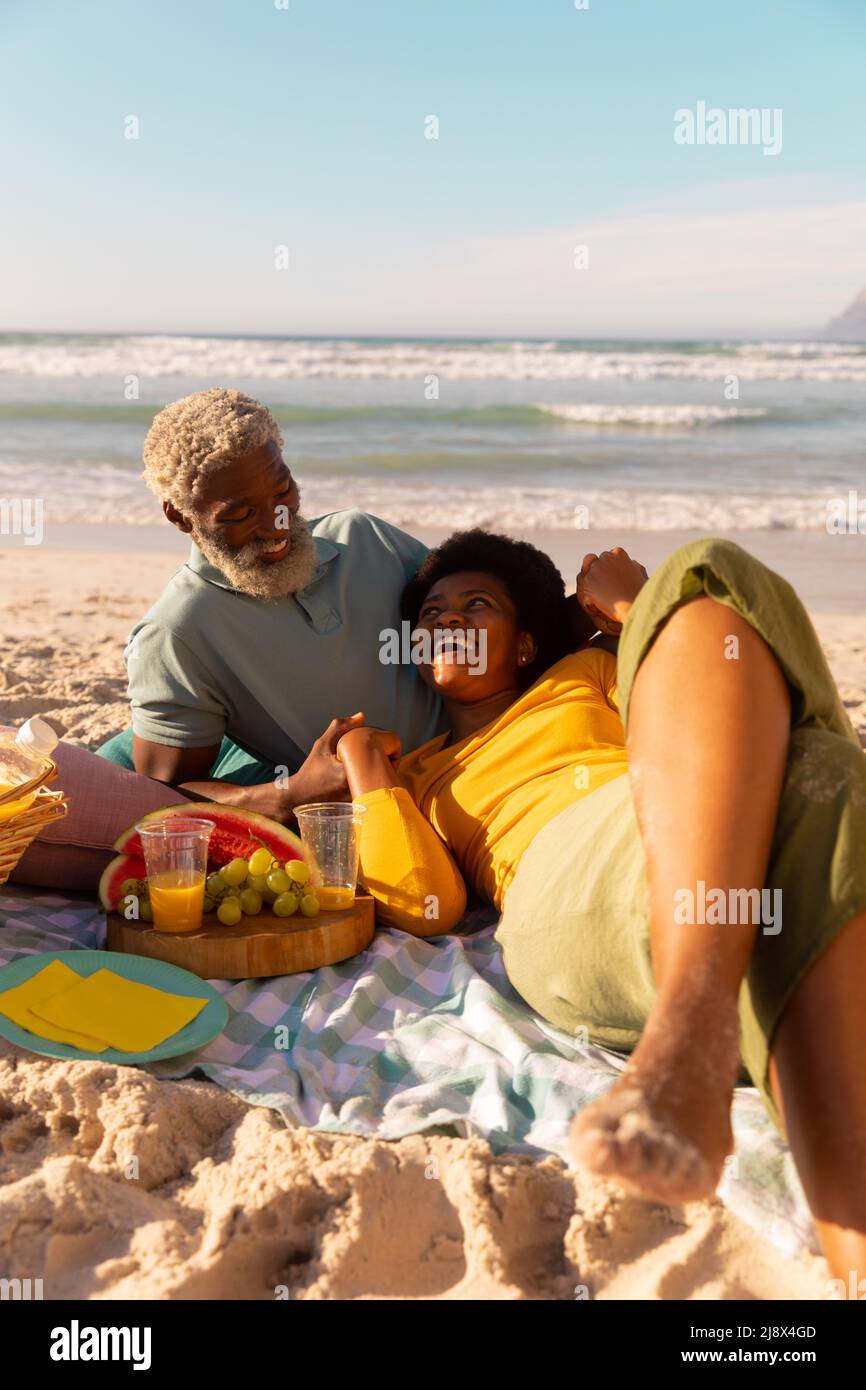 Donna matura afroamericana sdraiata con cibo e bevande sul braccio dell'uomo anziano contro il mare e il cielo limpido Foto Stock