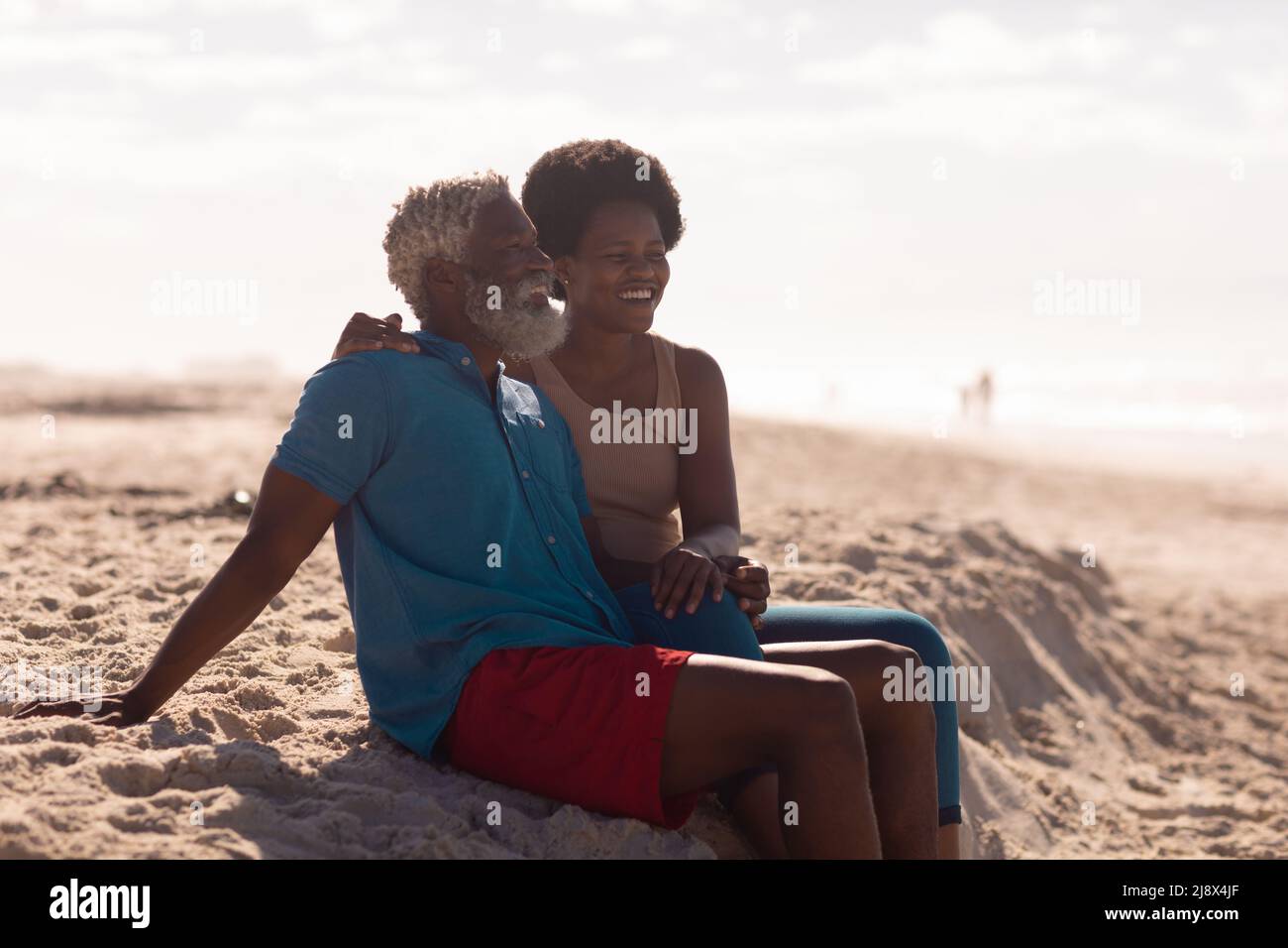 African american ported uomo anziano e donna matura con capelli afro seduti sulla spiaggia contro il cielo Foto Stock
