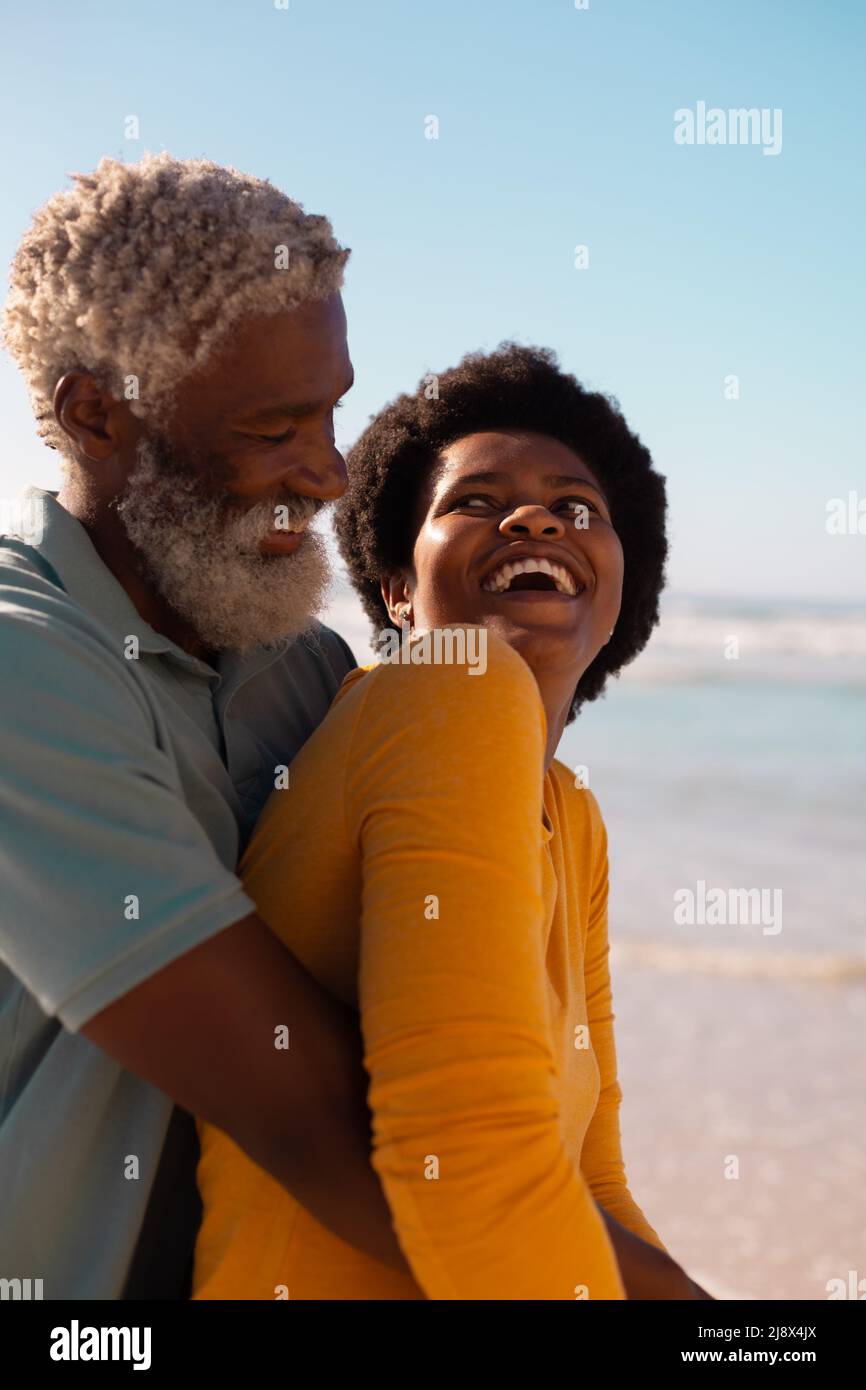 Uomo anziano afroamericano bearded che abbraccia la donna matura con i capelli afro alla spiaggia contro il cielo chiaro Foto Stock