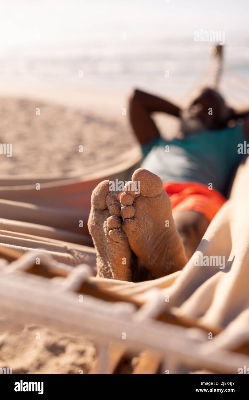 Uomo anziano afroamericano con sabbia sui piedi adagiato su amaca in spiaggia in giorno di sole Foto Stock
