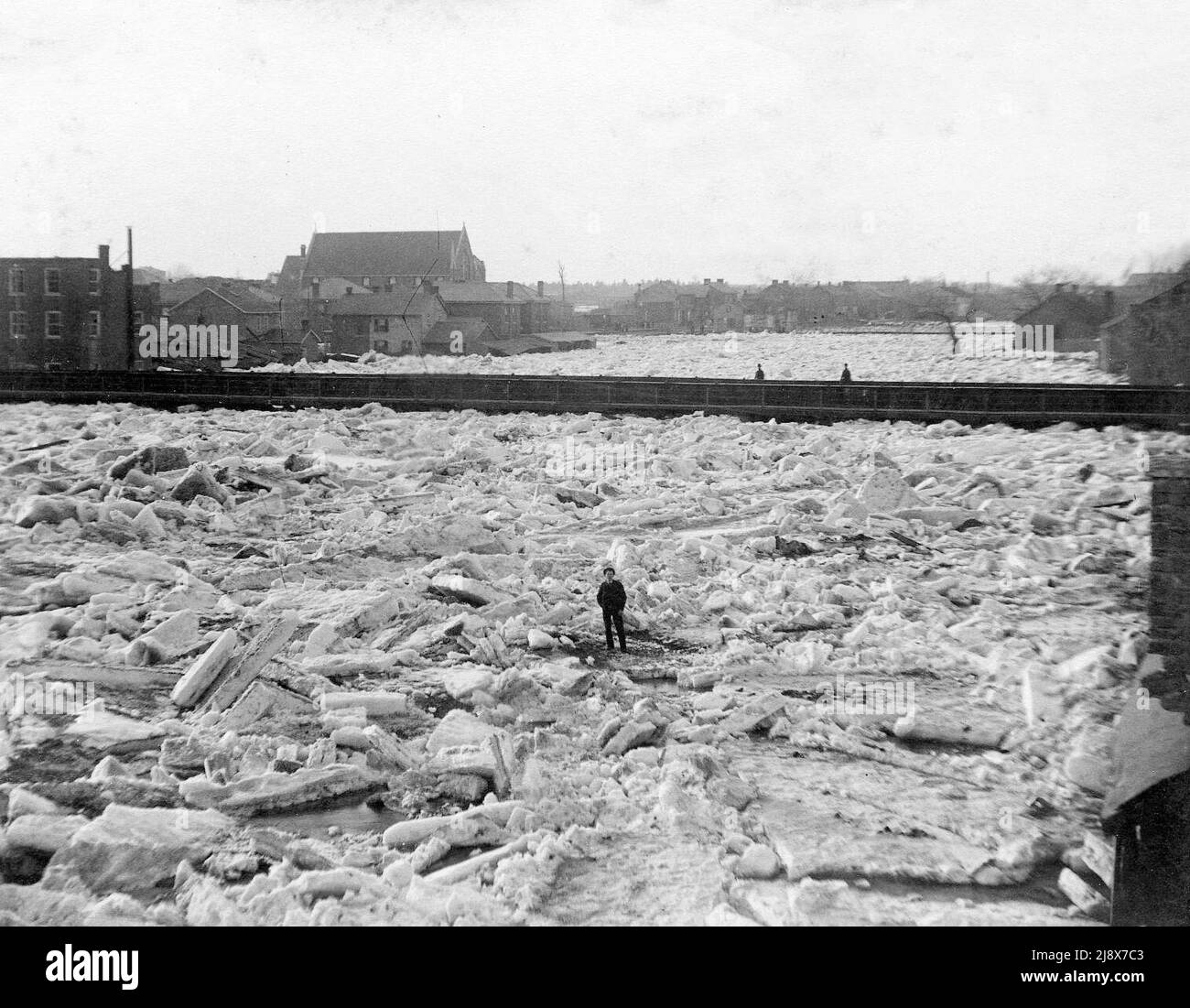 Persona in piedi sul ghiaccio sul fiume Moira a Belleville, Ontario durante l'alluvione del 1886, guardando a nord verso il ponte inferiore con la Chiesa di Cristo a metà sinistra e il ponte pedonale oltre ca. 1886 Foto Stock