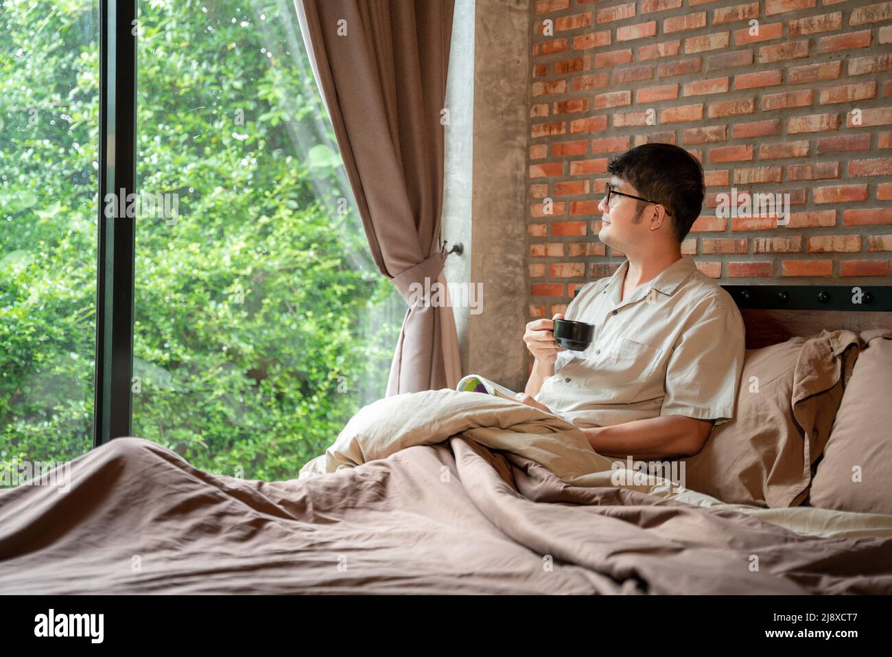 Un uomo sorride di buon umore al mattino con una tazza di bevanda calda caffè lettura libro in camera da letto a casa. Foto Stock