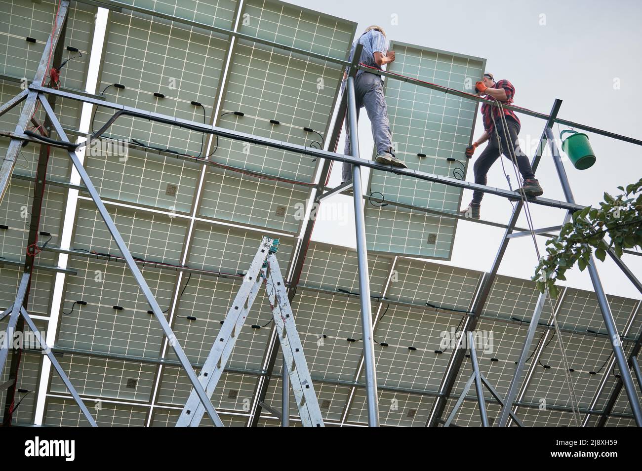 Lavoratori che installano pannelli solari su travi metalliche. Lavoro di squadra. Concetto di energia rinnovabile ed ecologica. Tecnologia moderna e innovazione. Idea di ambiente sicuro. Vista dal basso. Giorno Foto Stock