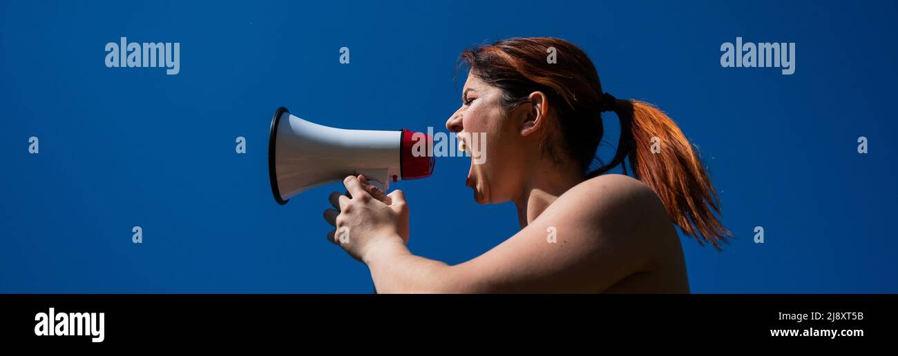 Donna caucasica dai capelli rossi urlando in un megafono contro un cielo blu Foto Stock