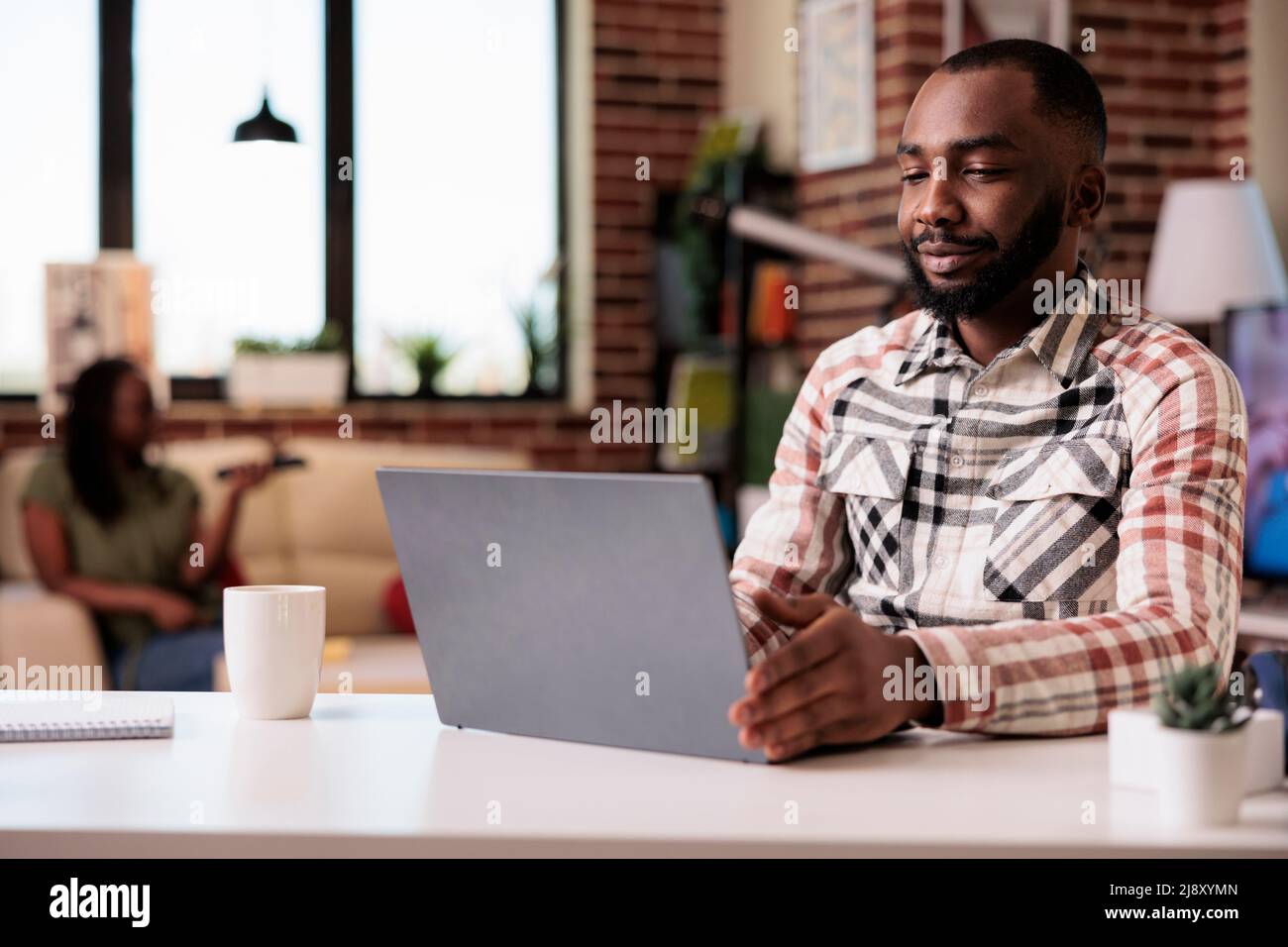 Ritratto di studente afroamericano rilassarsi guardando i contenuti video sul portatile mentre la ragazza si sta rilassando sul divano. Libera professionista maschile che utilizza il computer portatile streaming online show in salotto. Foto Stock