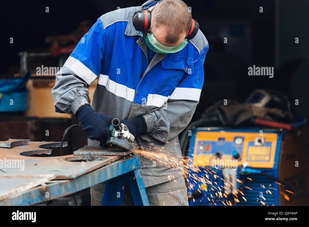 Il vero lavoratore in abiti da lavoro gratta la superficie metallica e le scintille volano. Ritratto reale della persona al lavoro Foto Stock
