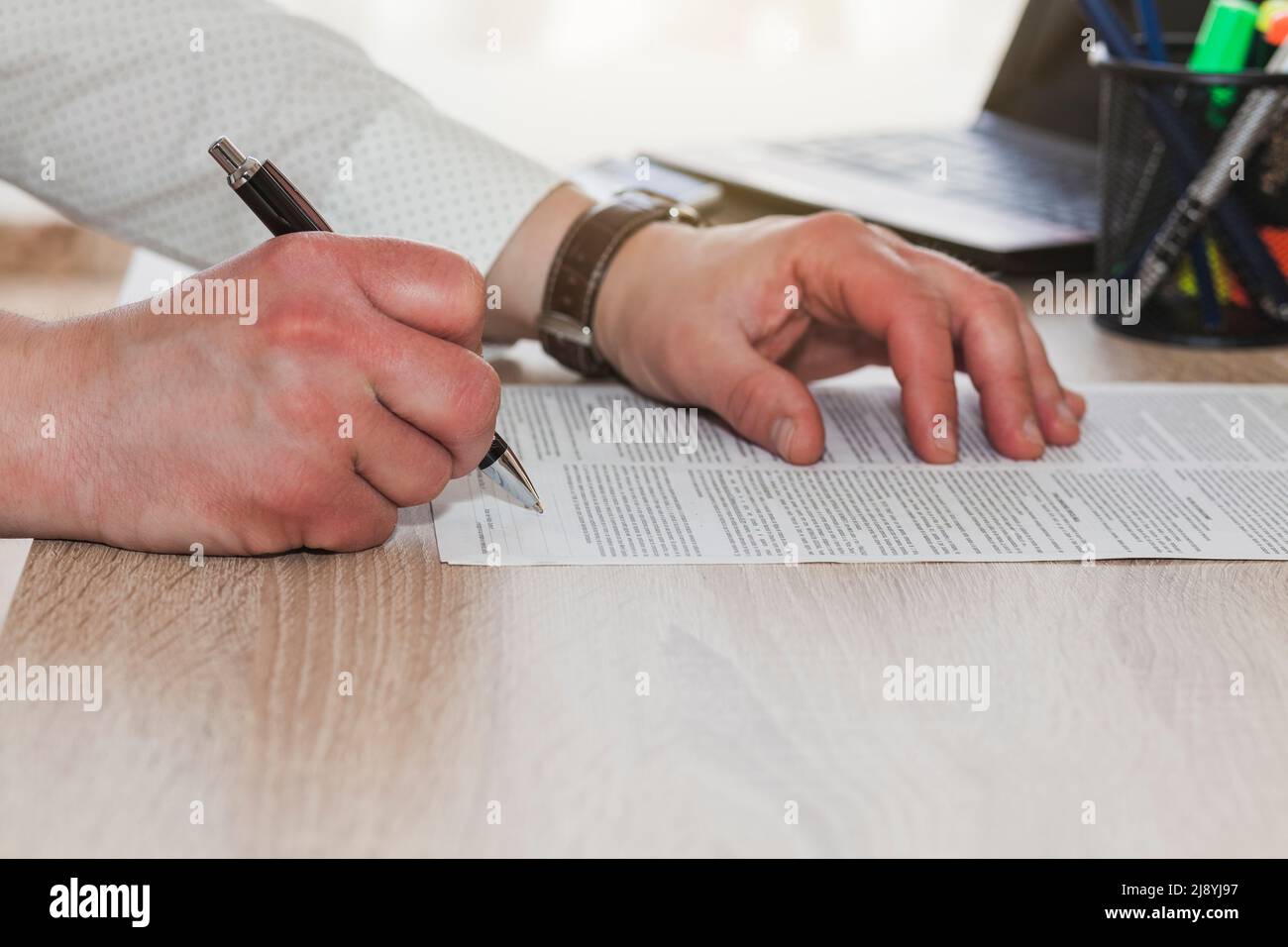 Le mani di un uomo sconosciuto che tiene una penna mentre si prepara a firmare un documento su un tavolo di legno. Diversi elementi dell'ufficio sono considerati fuori fuoco. Foto Stock
