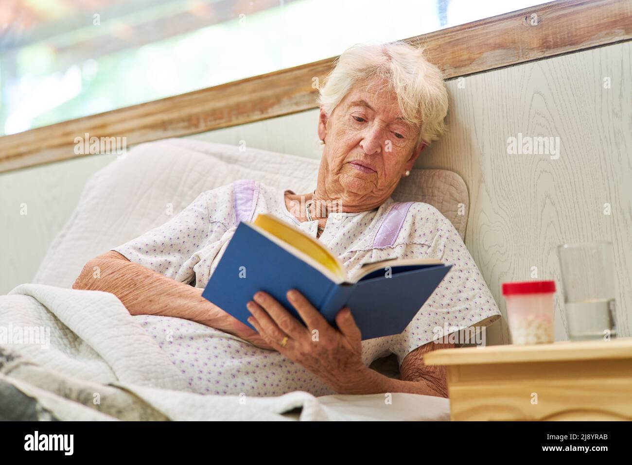 Donna anziana malata che si trova a letto leggendo un libro in ospedale o in casa di cura Foto Stock