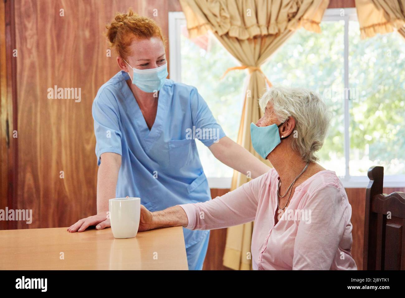 Donna anziana e infermiera geriatrica o infermiera con maschera facciale a causa di Covid-19 nella casa di cura Foto Stock