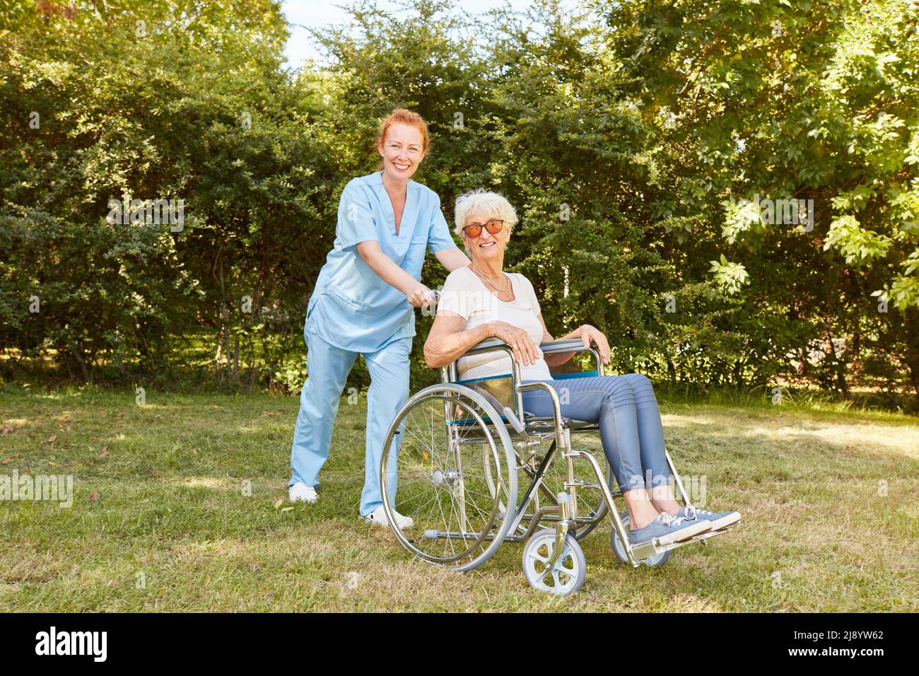 L'infermiera geriatrica spinge una donna anziana con disabilità in una sedia a rotelle attraverso il prato della clinica di riabilitazione Foto Stock