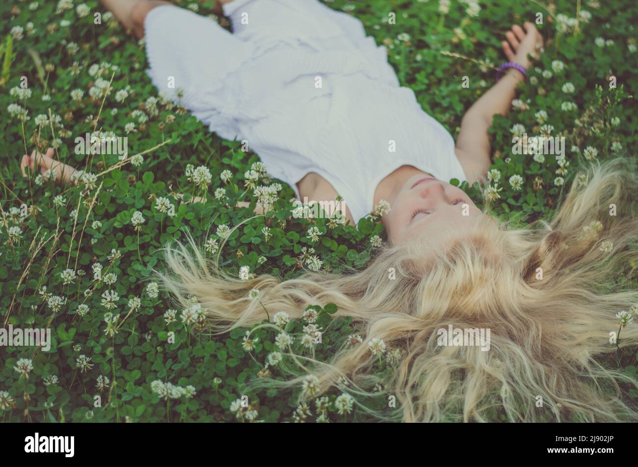 ragazza con capelli biondi lunghi giacenti in erba trifoglio verde con piccoli fiori bianchi, vista dalla prospettiva degli uccelli Foto Stock