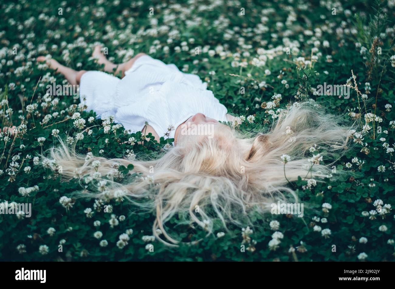 ragazza con capelli biondi lunghi giacenti in erba trifoglio verde con piccoli fiori bianchi, vista dalla prospettiva degli uccelli Foto Stock