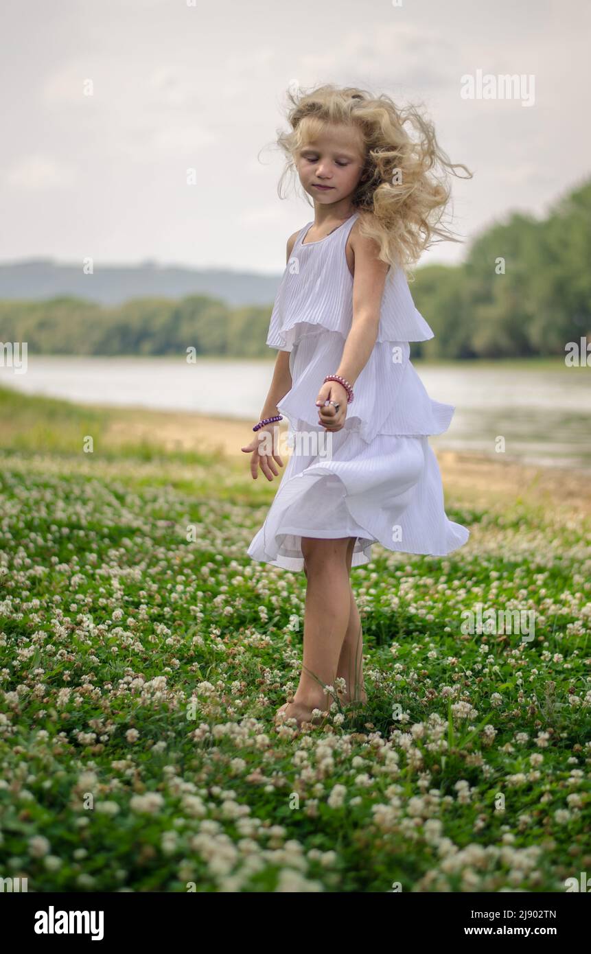 adorabile ragazza con capelli biondi lunghi e abito bianco che ballano in verde prato floreale Foto Stock