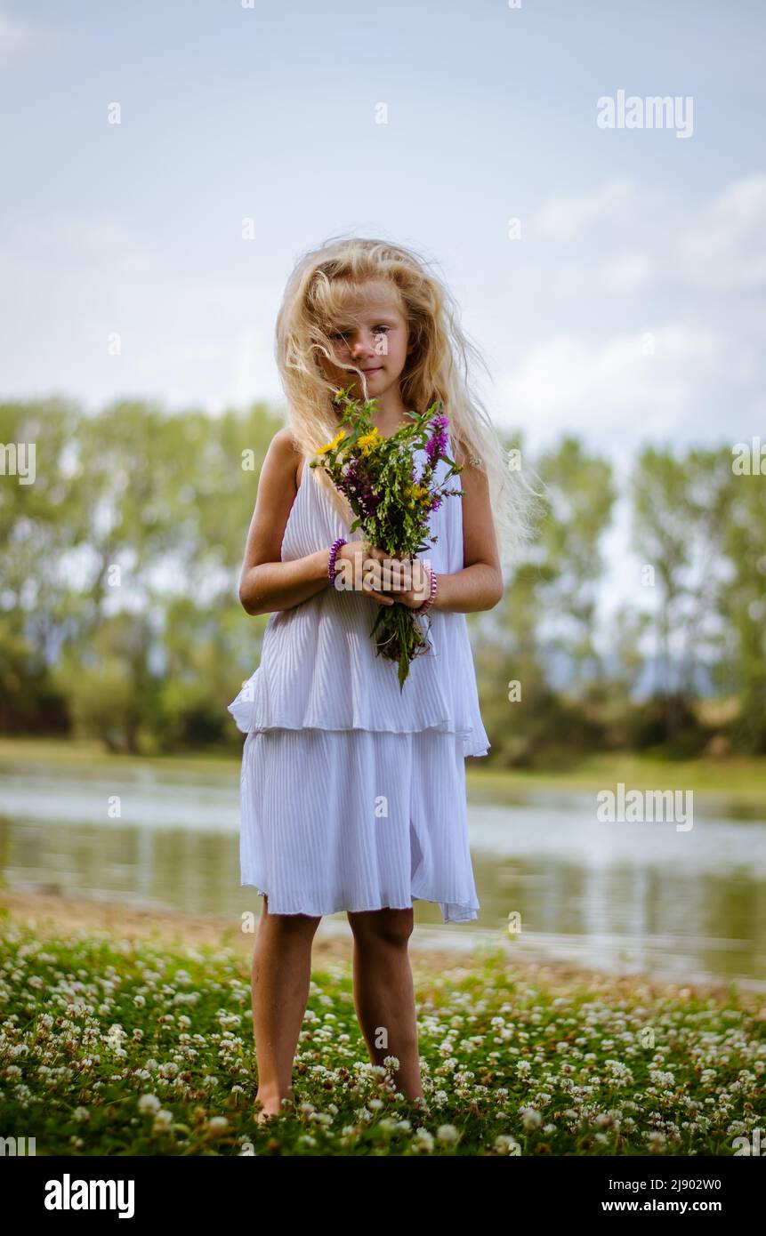 adorabile ragazza con capelli biondi lunghi e abito bianco che ballano in verde prato floreale Foto Stock