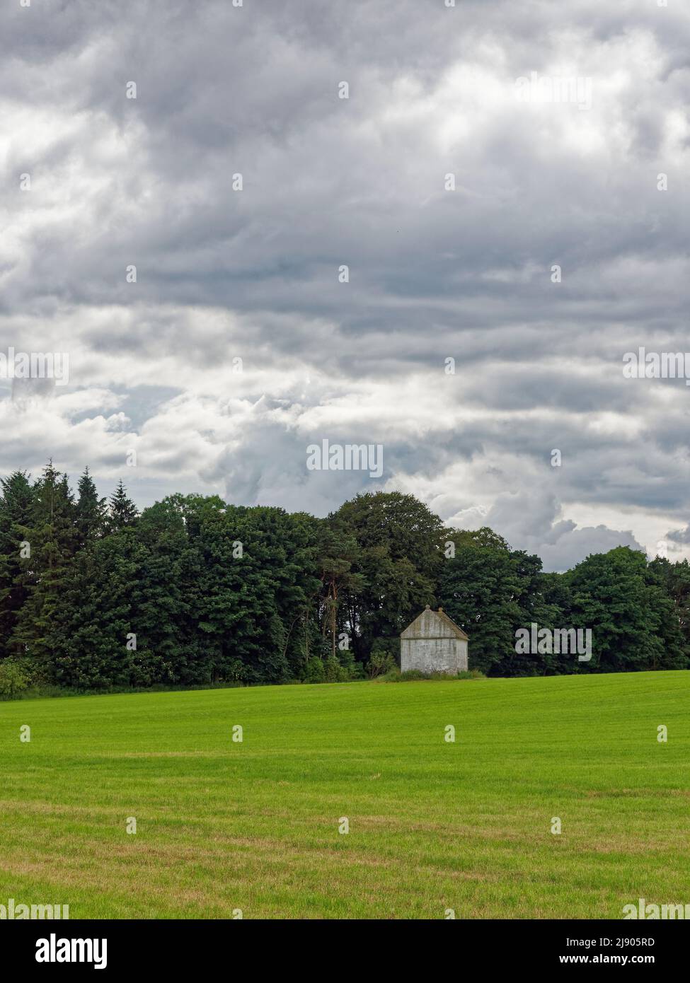 Un vecchio Doocot o Dovecote ben conservato situato in un campo di grano appena seminato con un bosco misto dietro e nuvole scure sopra. Foto Stock